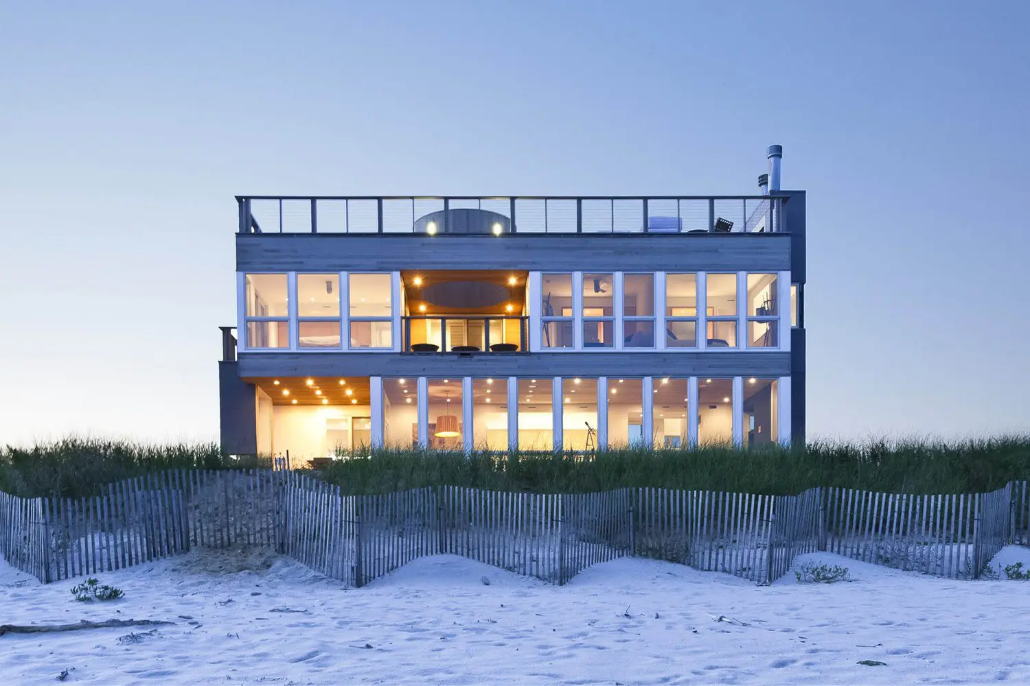 Modern beachfront house with large windows and balcony at dusk.