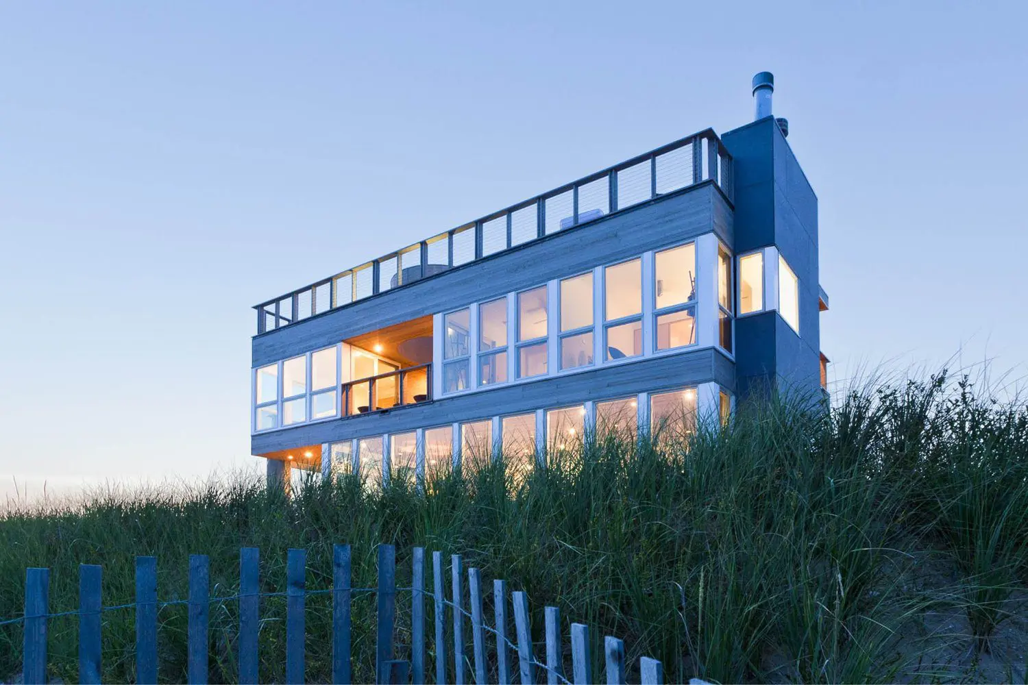 Modern glass house illuminated at dusk surrounded by greenery.