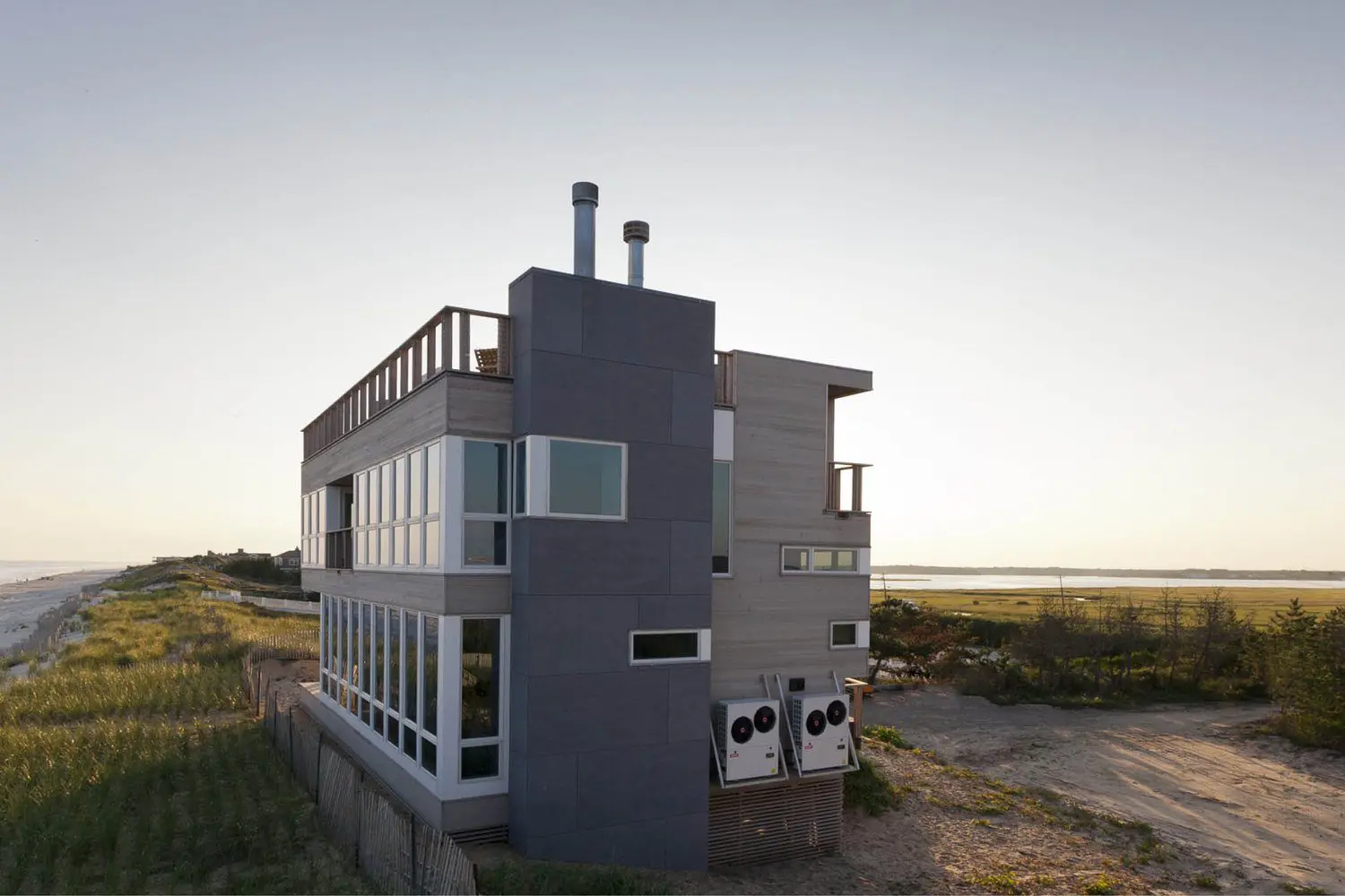 Modern gray beach house with large windows near the shore at sunset.