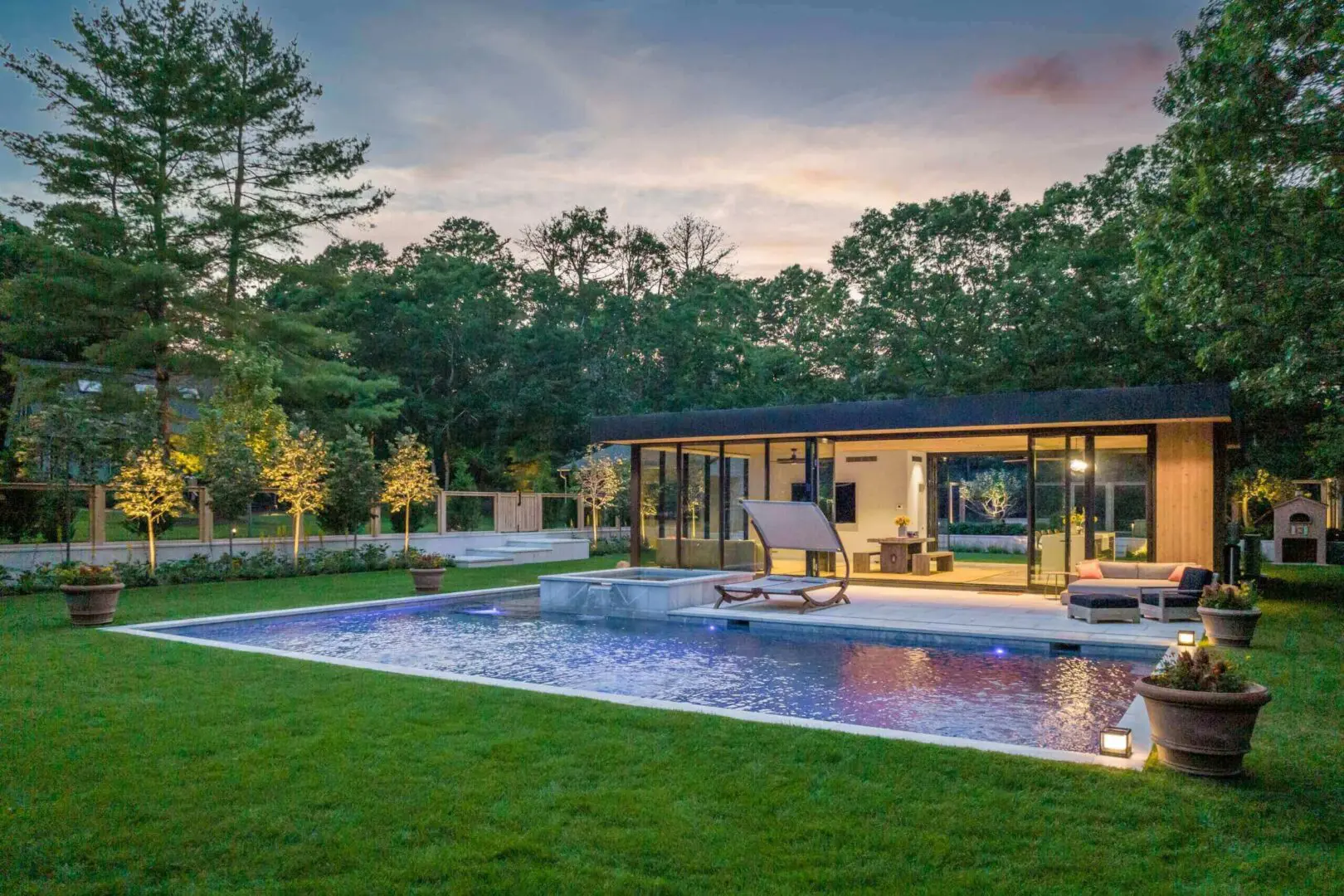 Modern backyard with a pool and illuminated pavilion at dusk.