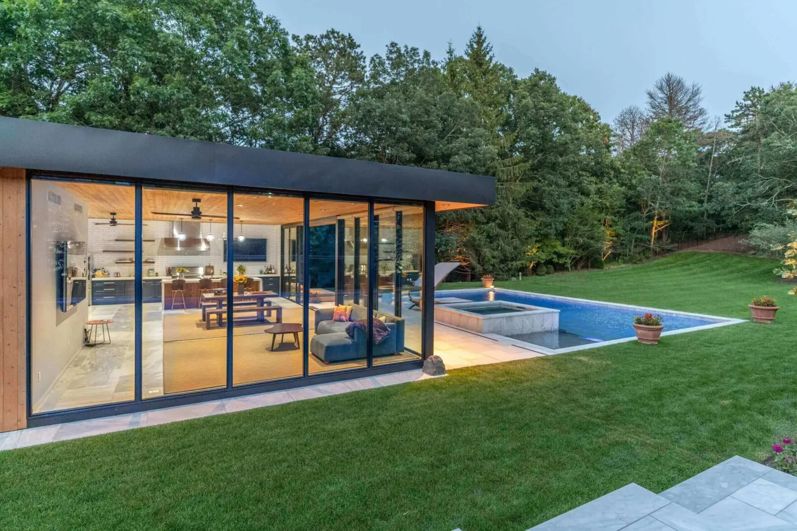Modern glass-walled house with a pool and lush green lawn at dusk.