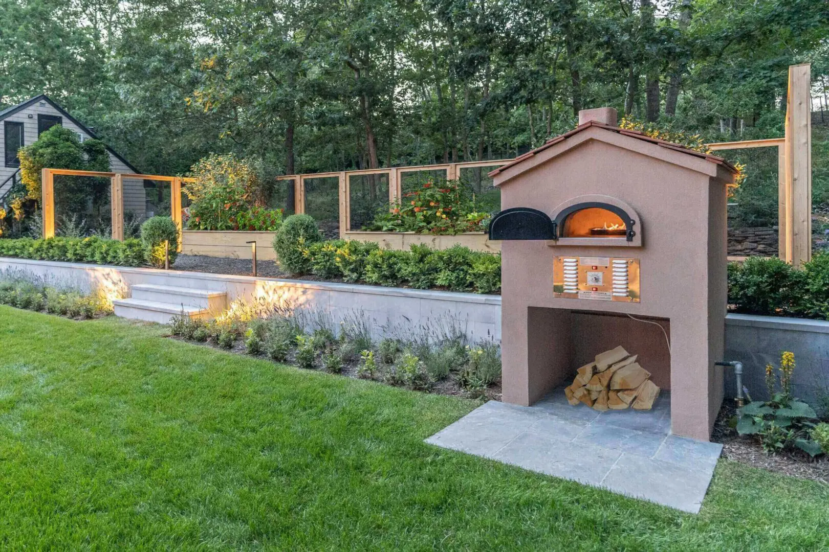 Outdoor wood-fired oven in a landscaped backyard with greenery and lighting.