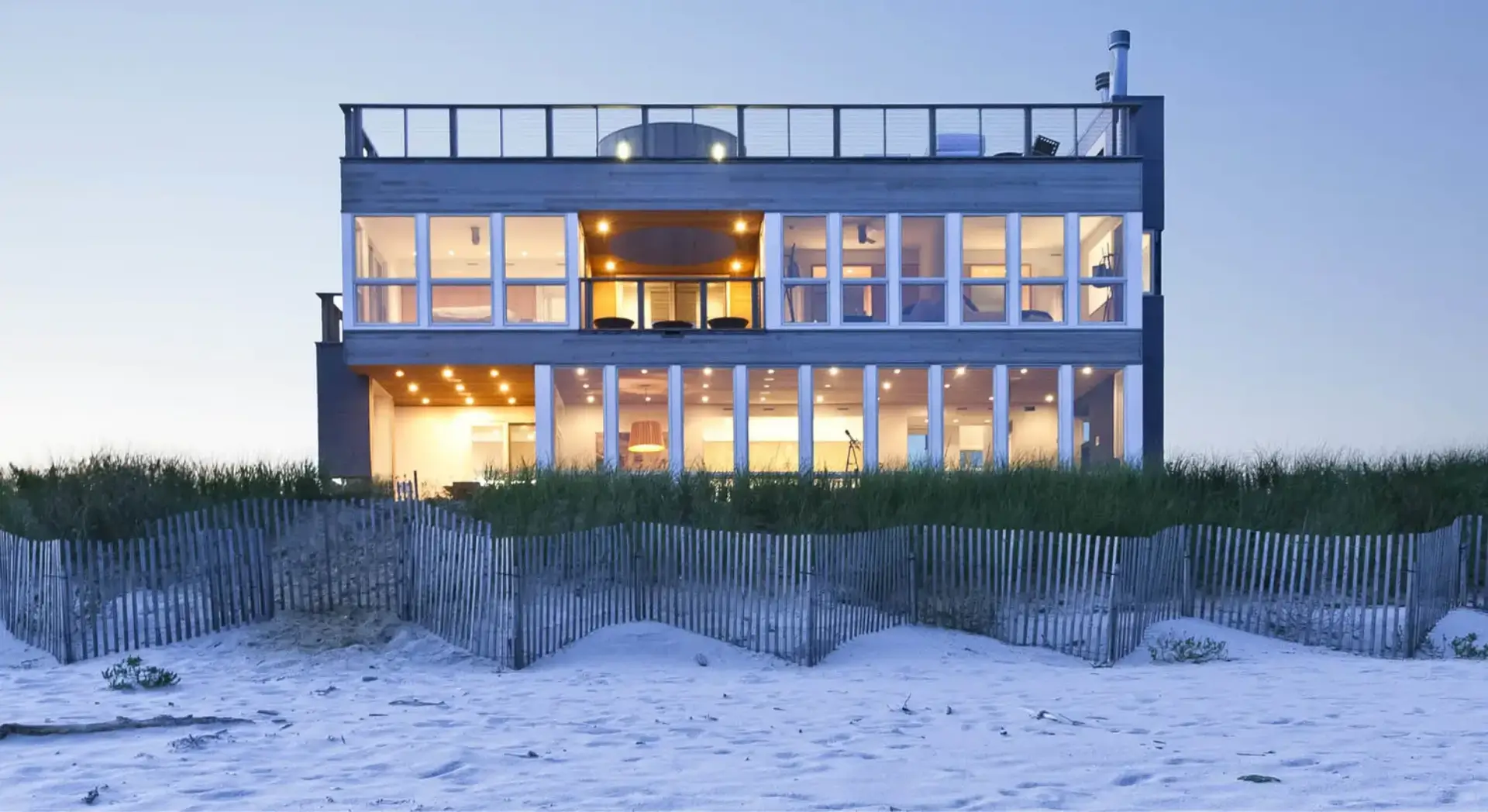 Modern beach house illuminated at dusk with large windows and wooden accents.